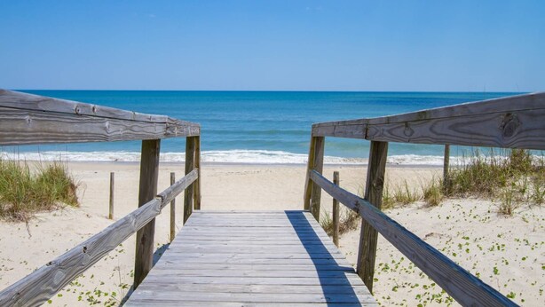 Een houten looppad dat naar een strand leidt, met helderblauw water en een zandstrand.