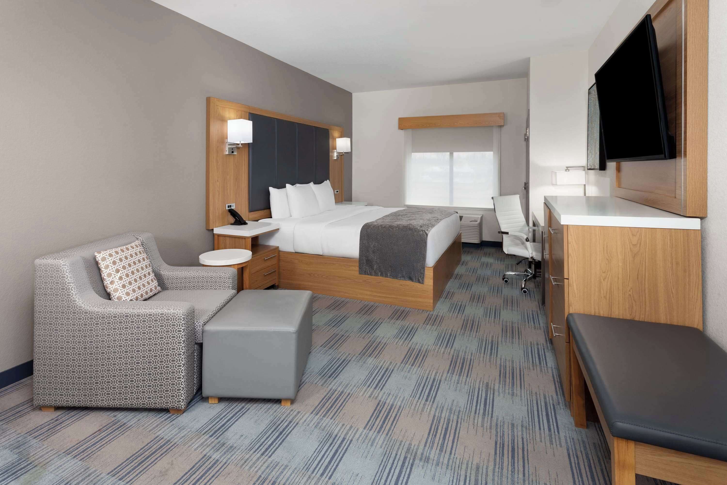 Una habitación de hotel moderna con una cama grande, una zona de estar con sofá y reposapiés, un escritorio y un televisor montado en la pared.