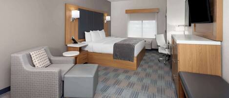 Una habitación de hotel moderna con una cama grande, una zona de estar con sofá y reposapiés, un escritorio y un televisor montado en la pared.