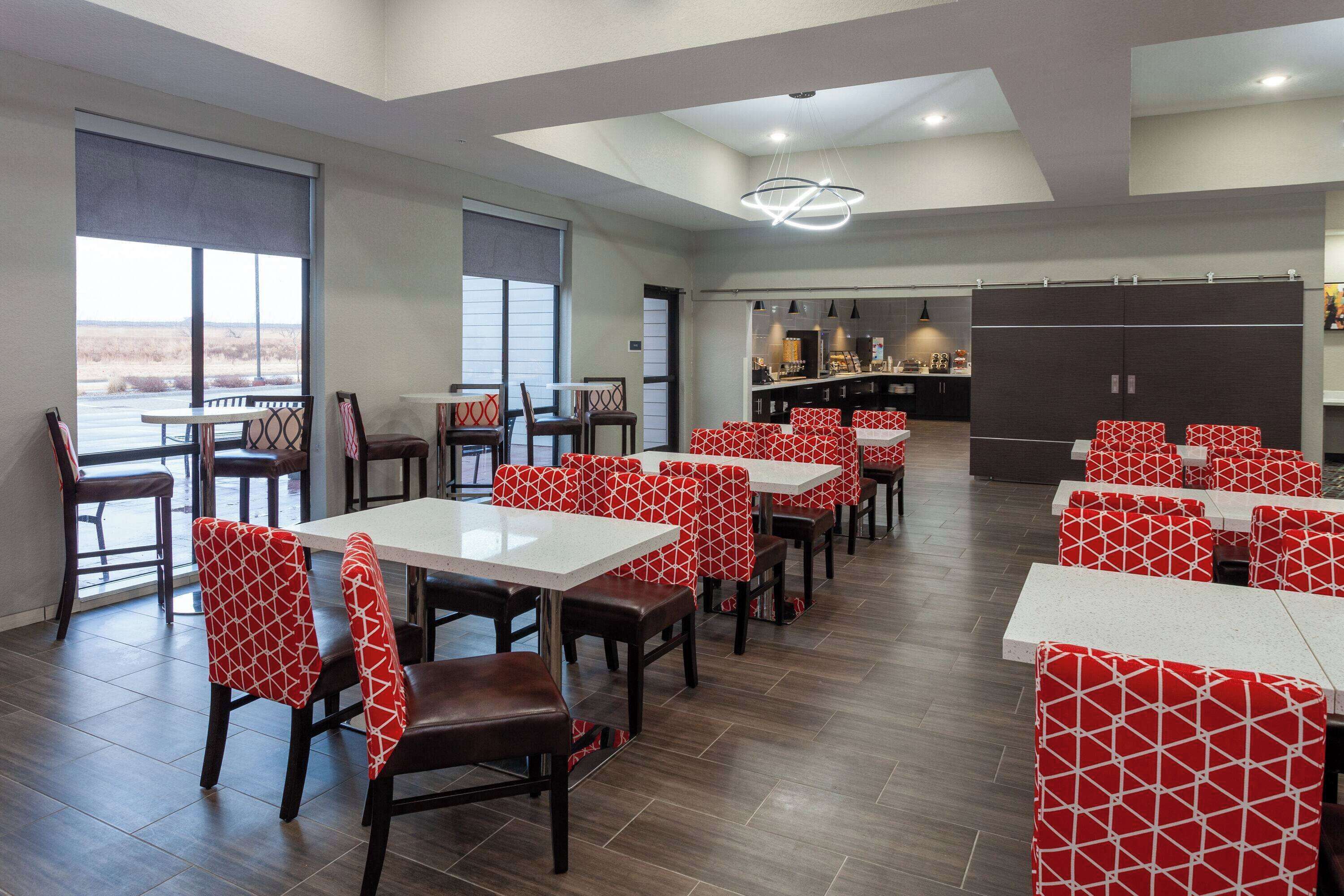 A dining area with tables and chairs, a kitchen in the background, and large windows.