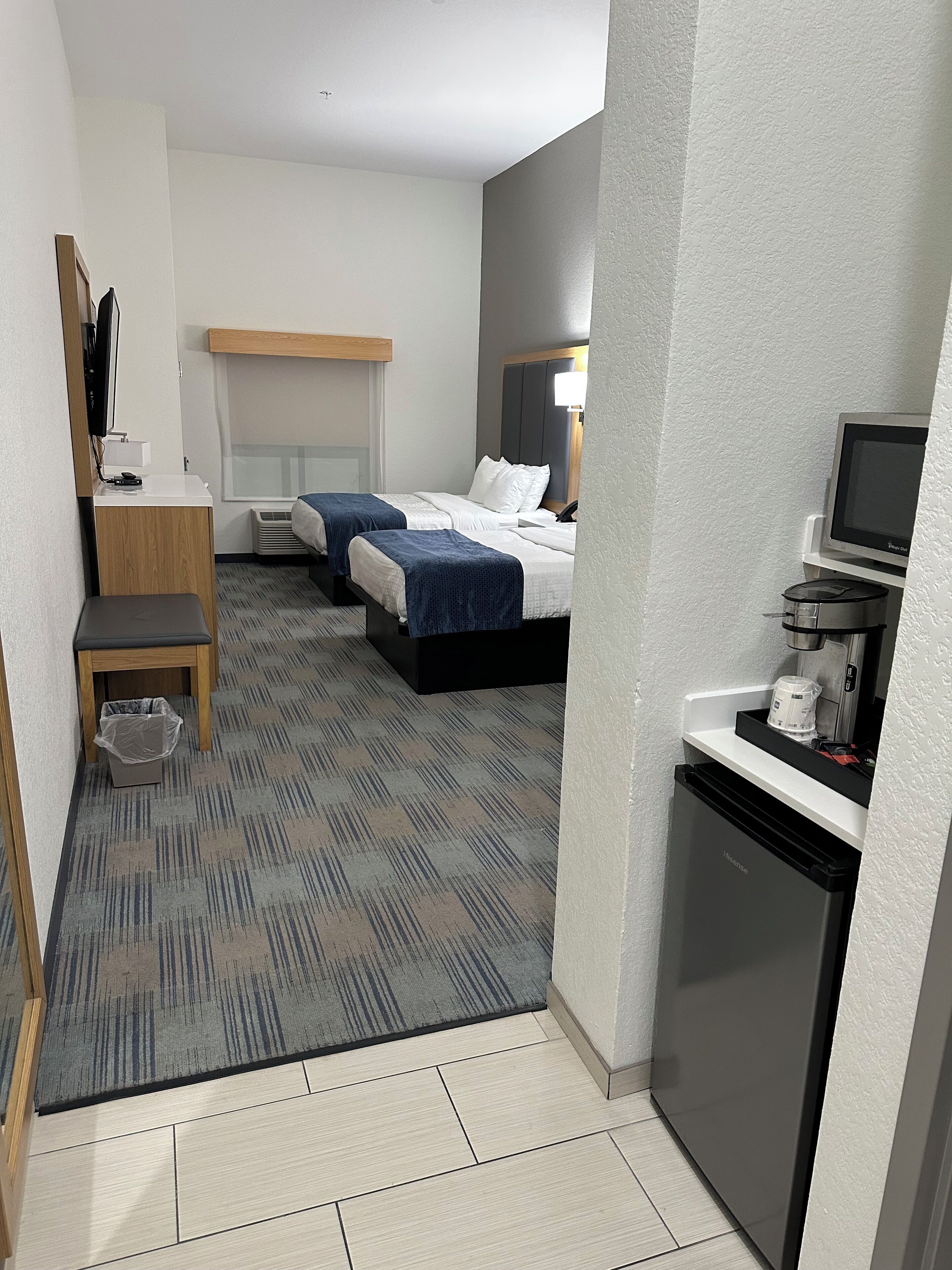 A hotel room with two beds, a desk, a small kitchenette, and a TV.