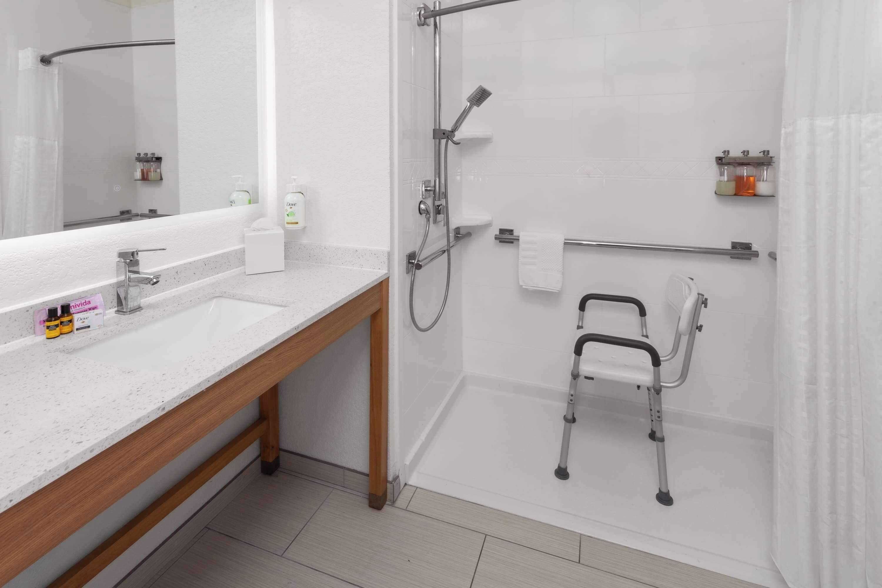 Un baño moderno con ducha, barra de apoyo, inodoro con barra de seguridad y tocador con espejo y lavamanos.