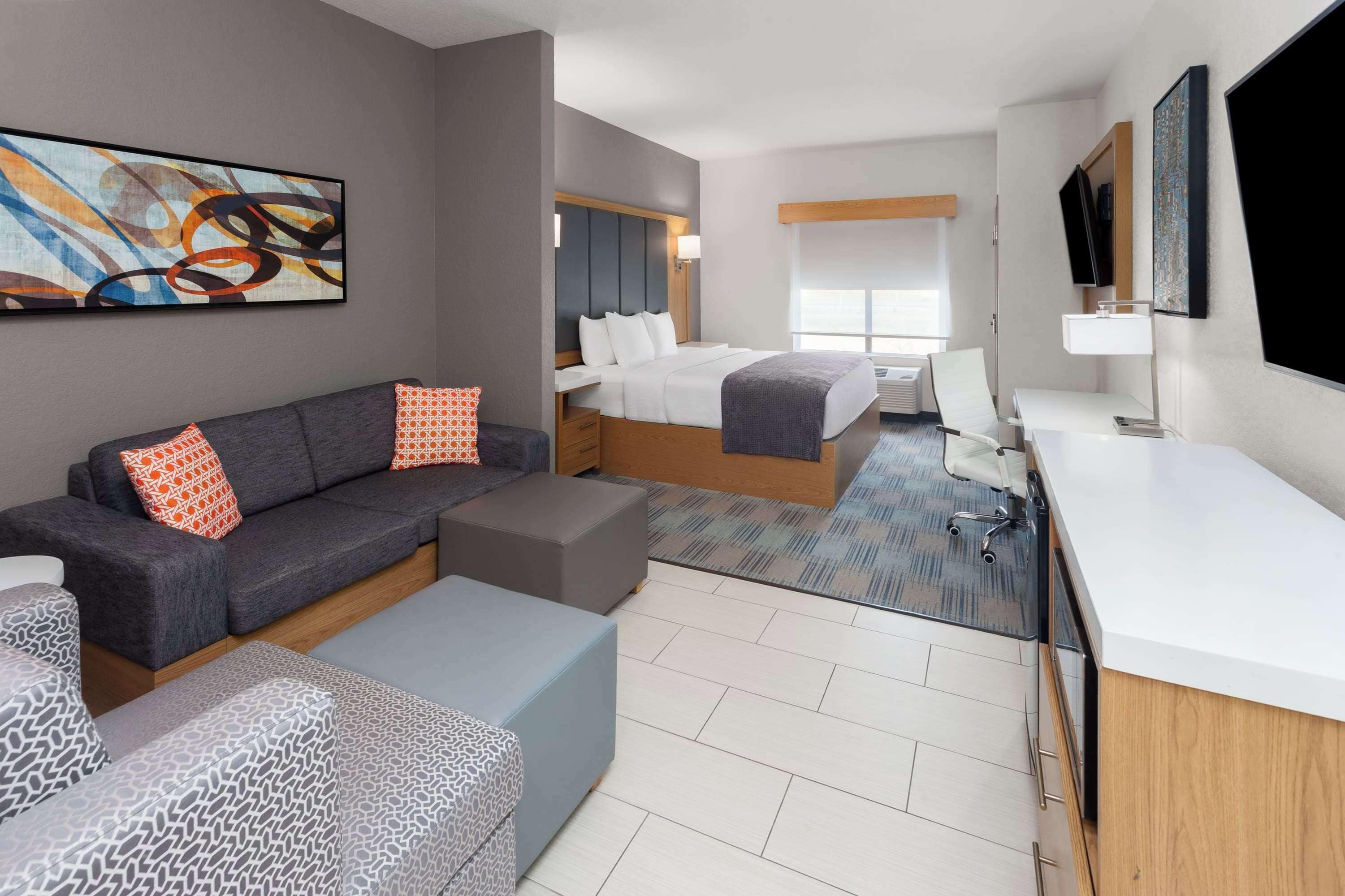 A modern hotel room with a gray sofa, a gray patterned ottoman, a bed with white linens, a desk with a white sink, and a large abstract painting on the wall.