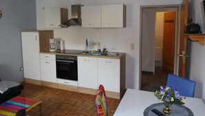 Een kleine keuken met witte kastjes, een fornuis en een spoelbak. Er staat een eettafel met een vaas bloemen.