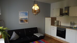 Een knus woongedeelte met een zwarte bank, een kleurrijke gestreepte tafel, een keuken met een kookplaat en oven, en een hanglamp.