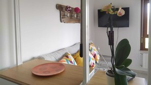 A modern living room with a wooden table, a sofa with colorful cushions, a flat-screen TV mounted on the wall, and a potted plant. - Monolocale La Fornacetta (Pisa)