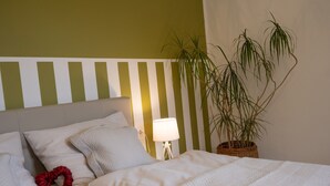 Una cama bien hecha con ropa de cama blanca y un adorno rojo, una lámpara de noche y una planta en maceta sobre una mesita.