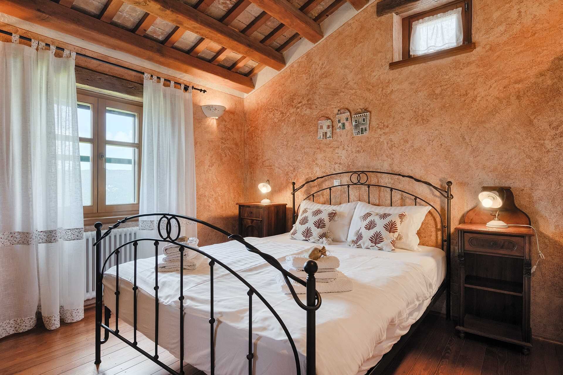 A bedroom with a metal bed frame, white bedding, a wooden floor, a window with curtains, and a small wall-mounted picture.