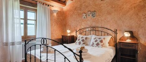 A bedroom with a metal bed frame, white bedding, a wooden floor, a window with curtains, and a small wall-mounted picture.