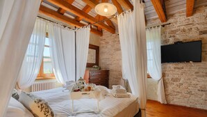 A bedroom with a wooden ceiling, stone walls, a bed with white bedding, a bedside table with a lamp, and a TV mounted on the wall.