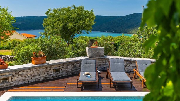 A poolside area with lounge chairs, a stone wall, and a view of a lake and mountains.