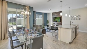 Een moderne keuken met een centraal eiland, een eethoek met een glazen tafel, en een woonruimte met een bank en televisie.