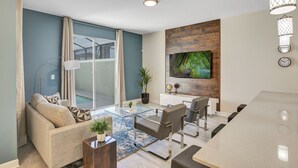 Een moderne woonkamer met een bank, fauteuils, een glazen salontafel, een tv bevestigd aan een houten wand en een schuifdeur van glas die leidt naar een balkon.