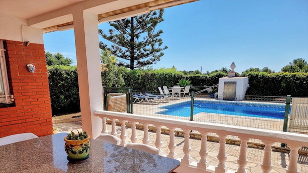 Un patio cubierto con piscina, sillas blancas y una mesa con planta en maceta.