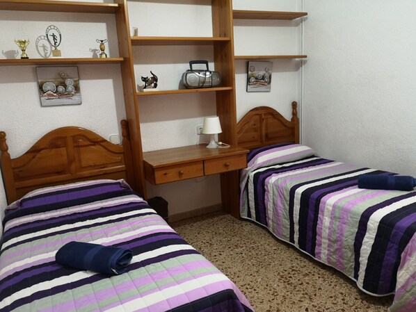A bedroom with two beds, wooden headboards, a desk with a lamp, and shelves with various items.