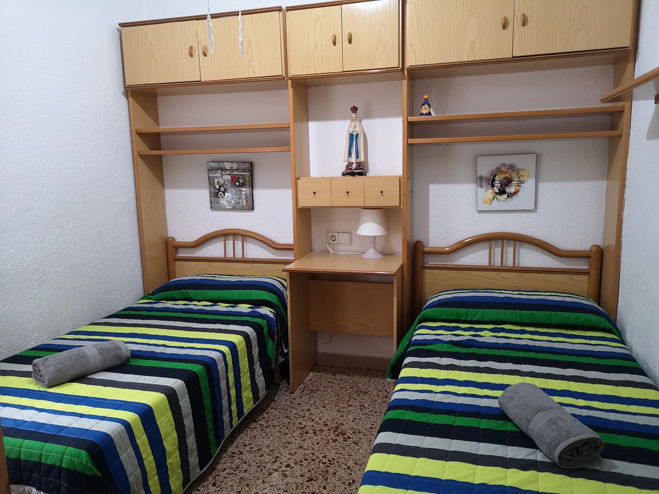 Une chambre avec deux lits simples, des armoires en bois, un bureau et une petite étagère ornée d’objets décoratifs.