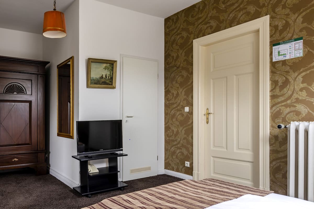 A hotel room with a wooden wardrobe, a television on a black stand, a patterned wallpaper, a white door, and a radiator.