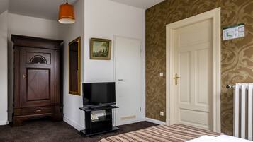 A hotel room with a wooden wardrobe, a television on a black stand, a patterned wallpaper, a white door, and a radiator.