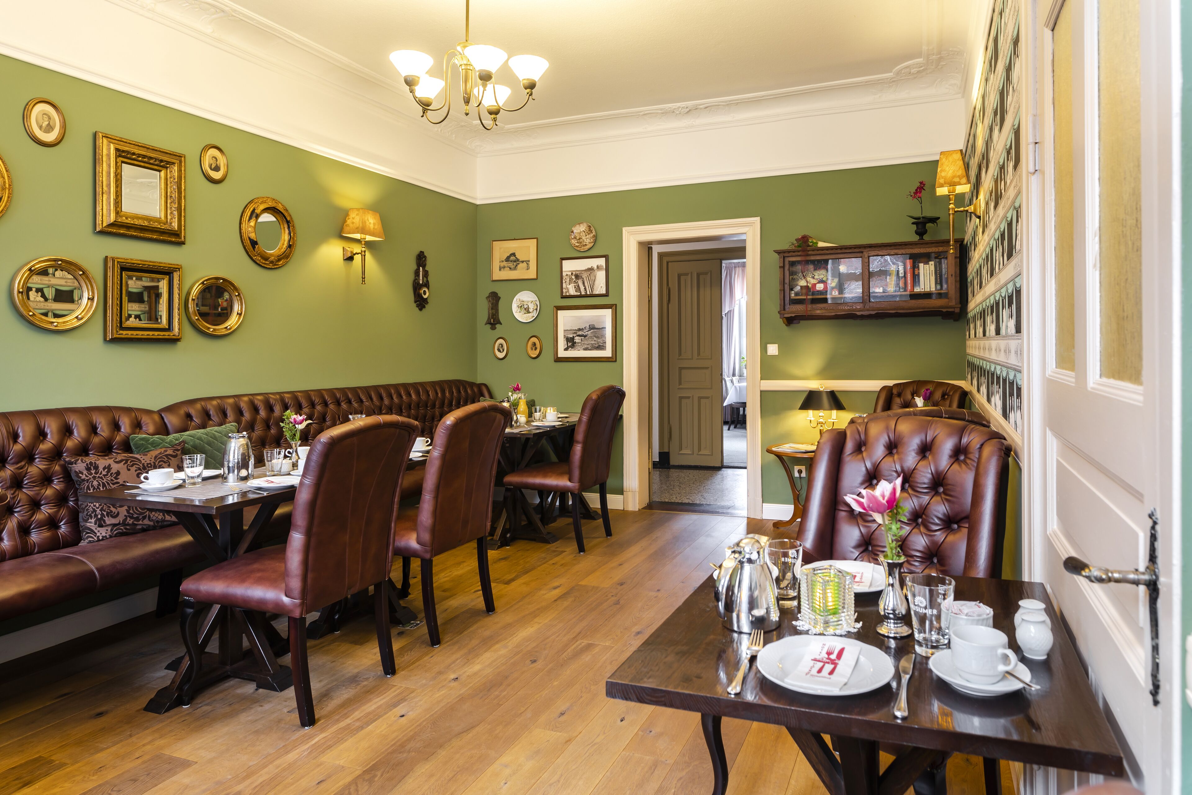 A dining area with a wooden table set for four, surrounded by leather chairs, a green wall with framed pictures, and a chandelier.