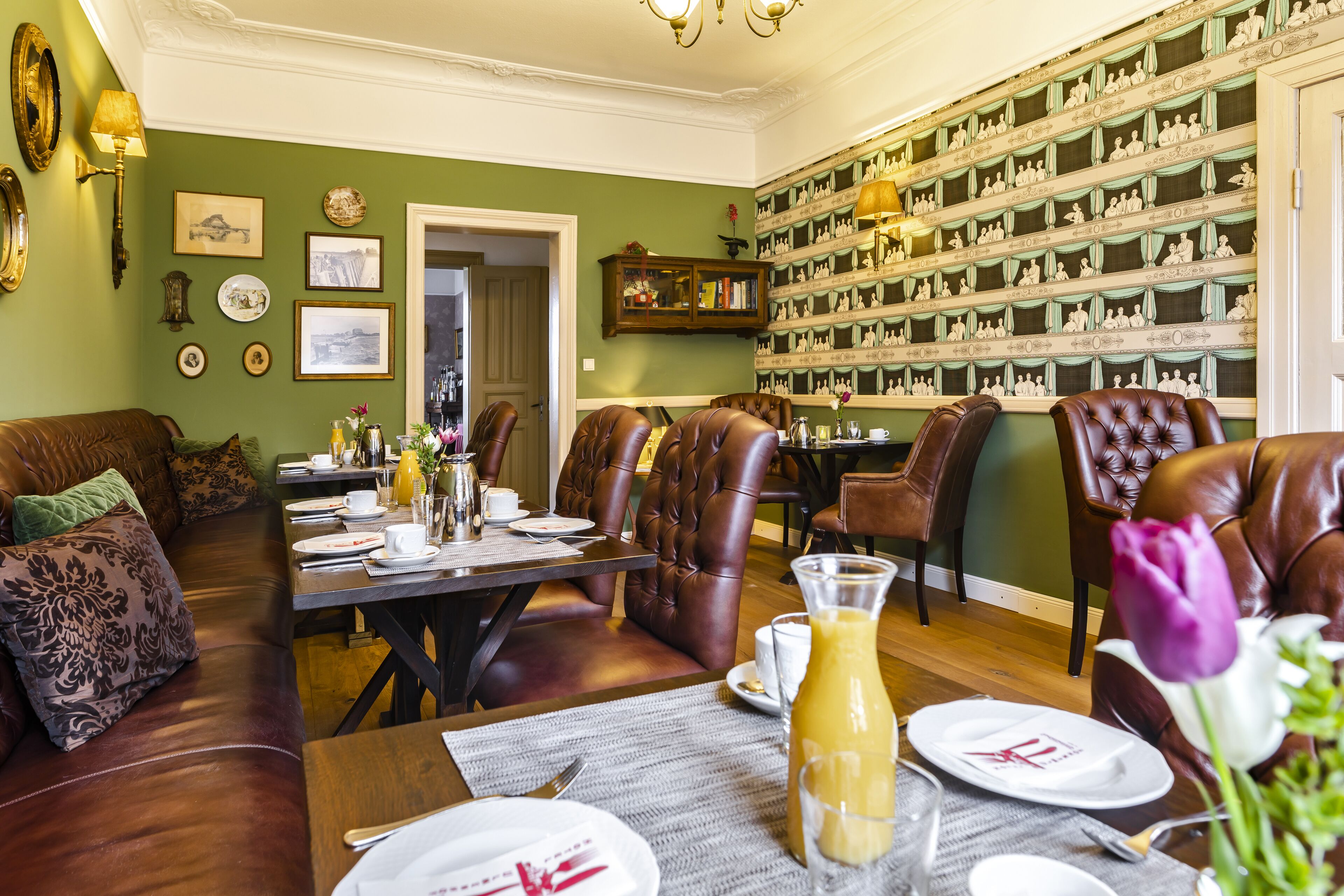 A dining area with a table set for four, surrounded by leather chairs, a bookshelf with decorative items, and a green wall with framed pictures.