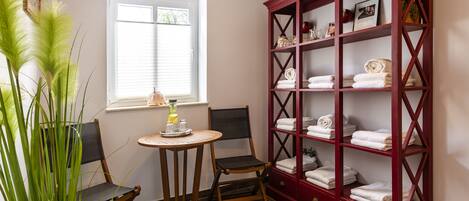A room with a wooden chair, a small round table with a tray, a red wooden cabinet with towels, and a window with blinds.