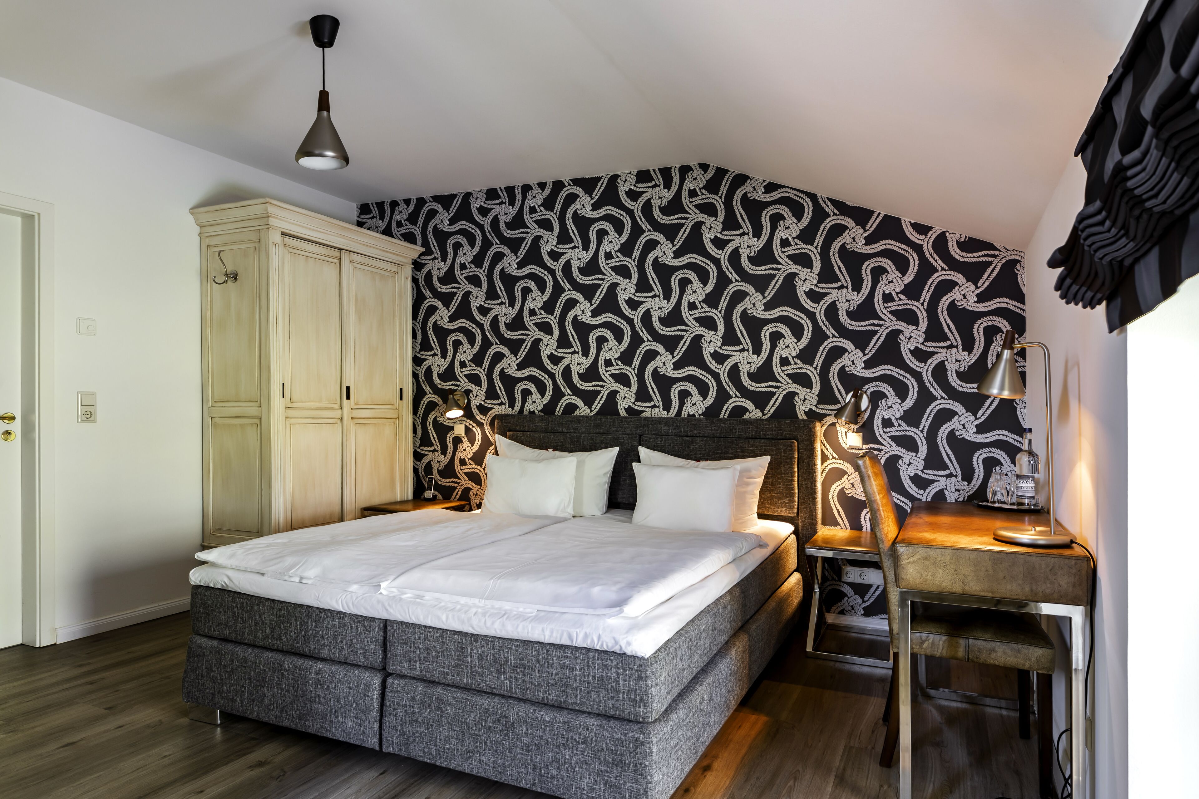 A bedroom with a bed, a desk, and a chair. There is a wardrobe and a patterned wall.