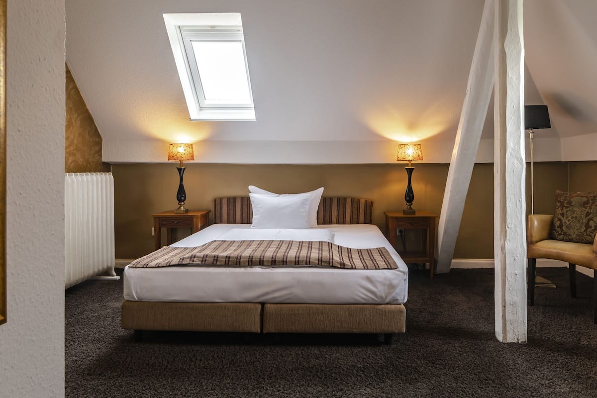 A bedroom with a skylight, two bedside lamps, a bed with a striped blanket, a chair, and a radiator.