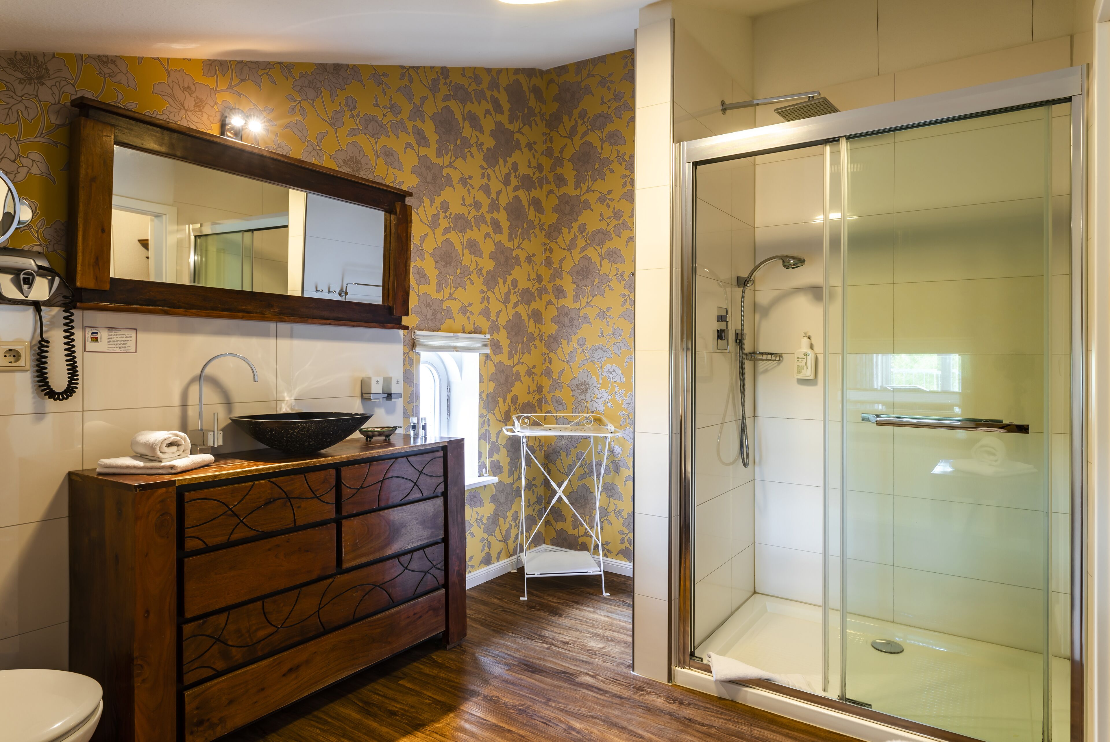 A bathroom with a wooden vanity, a mirror, a sink, a shower, and a tiled floor.