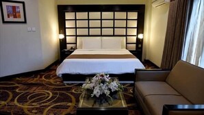 A hotel room with a large bed, a sofa, a glass coffee table with flowers, and a patterned carpet. - Hotel One Sukkur (Sukkur)