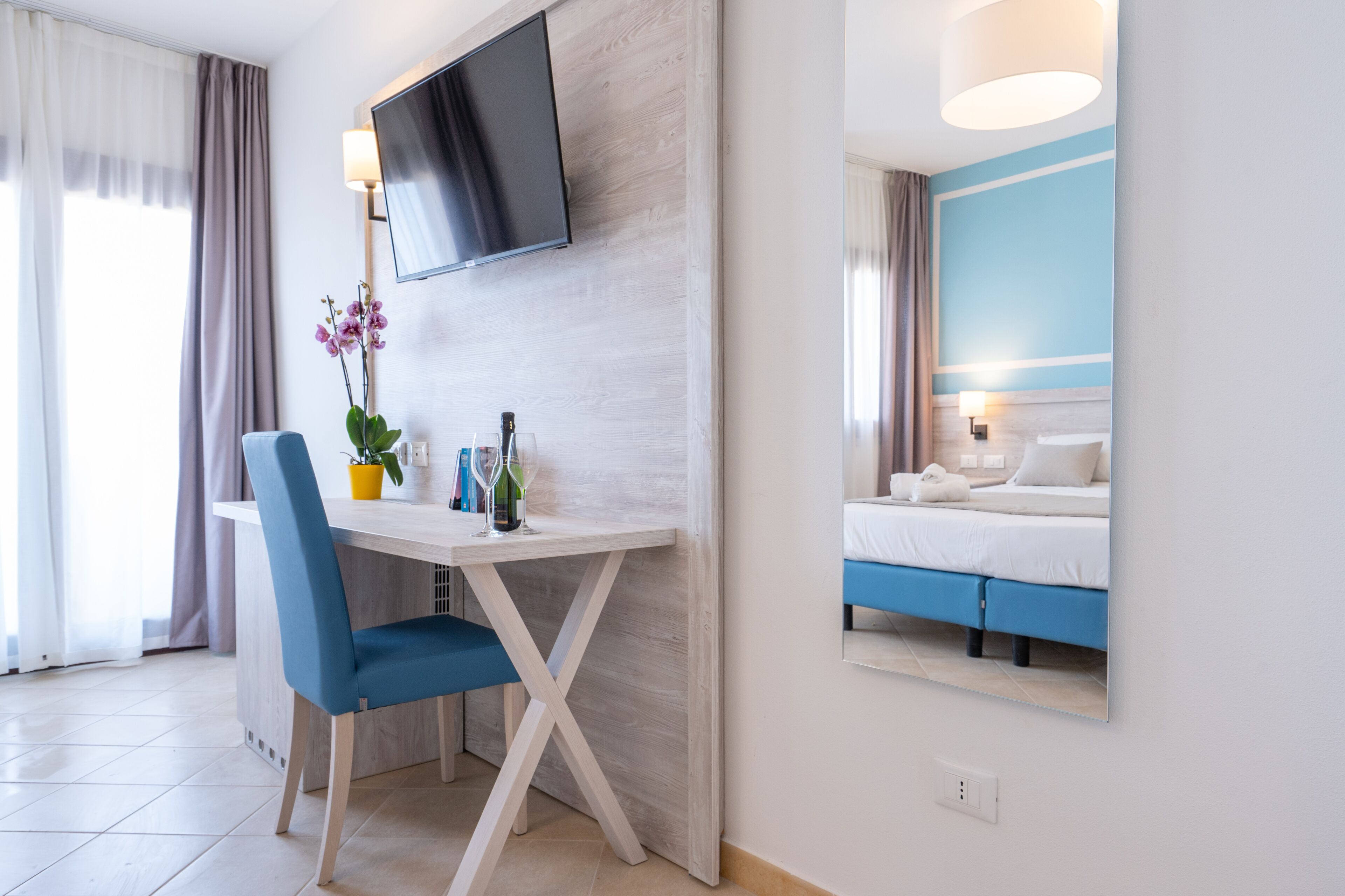 Ein Hotelzimmer mit einem Bett, einem Schreibtisch mit Stuhl, einem an der Wand montierten Fernseher und einem Fenster mit Vorhängen.