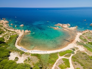Vista aérea de una zona costera con aguas turquesas cristalinas, afloramientos rocosos y una playa arenosa con sombrillas.