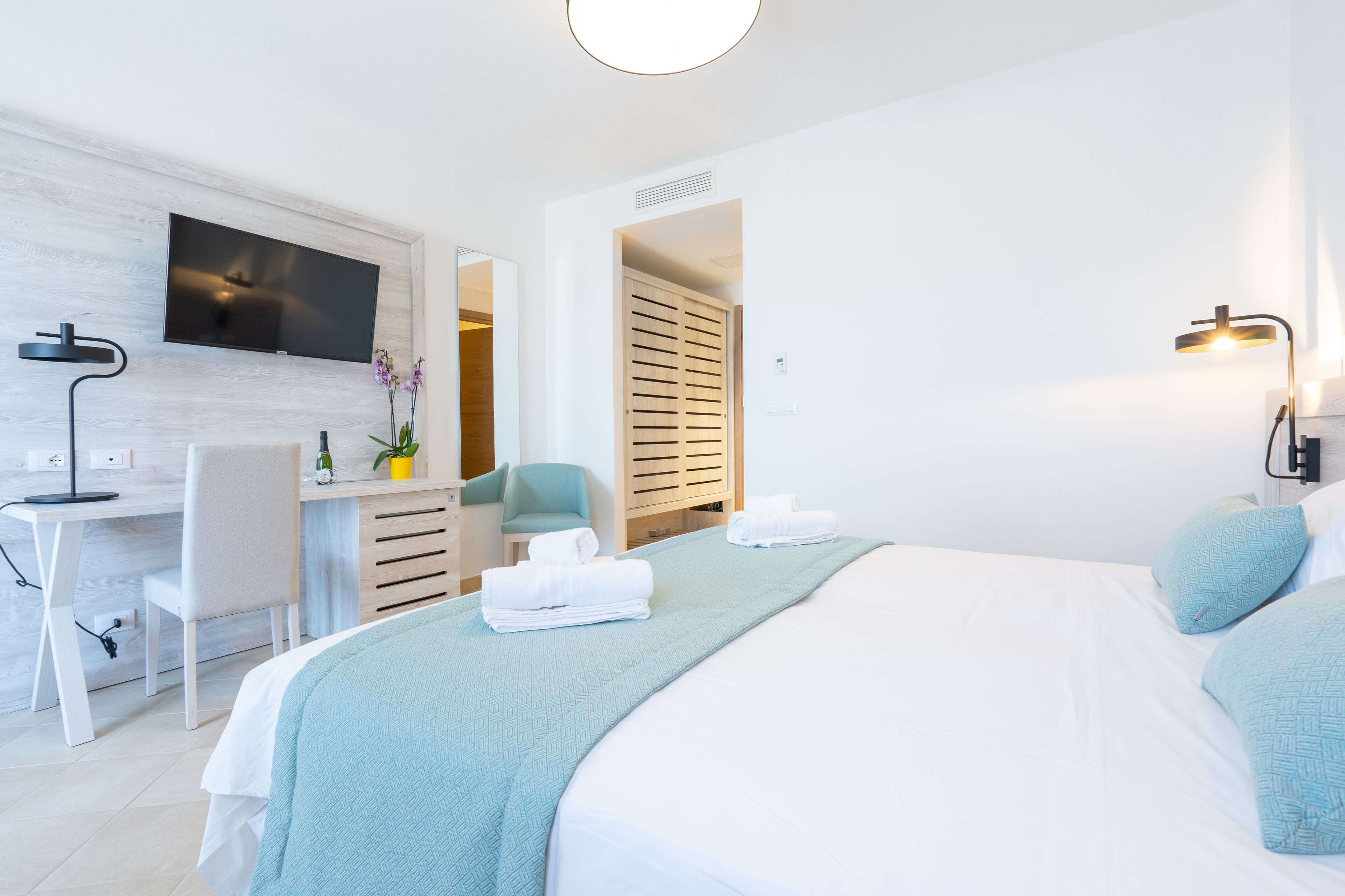 A modern hotel room with a bed, a desk, a chair, a TV, and a bathroom visible through an open door.