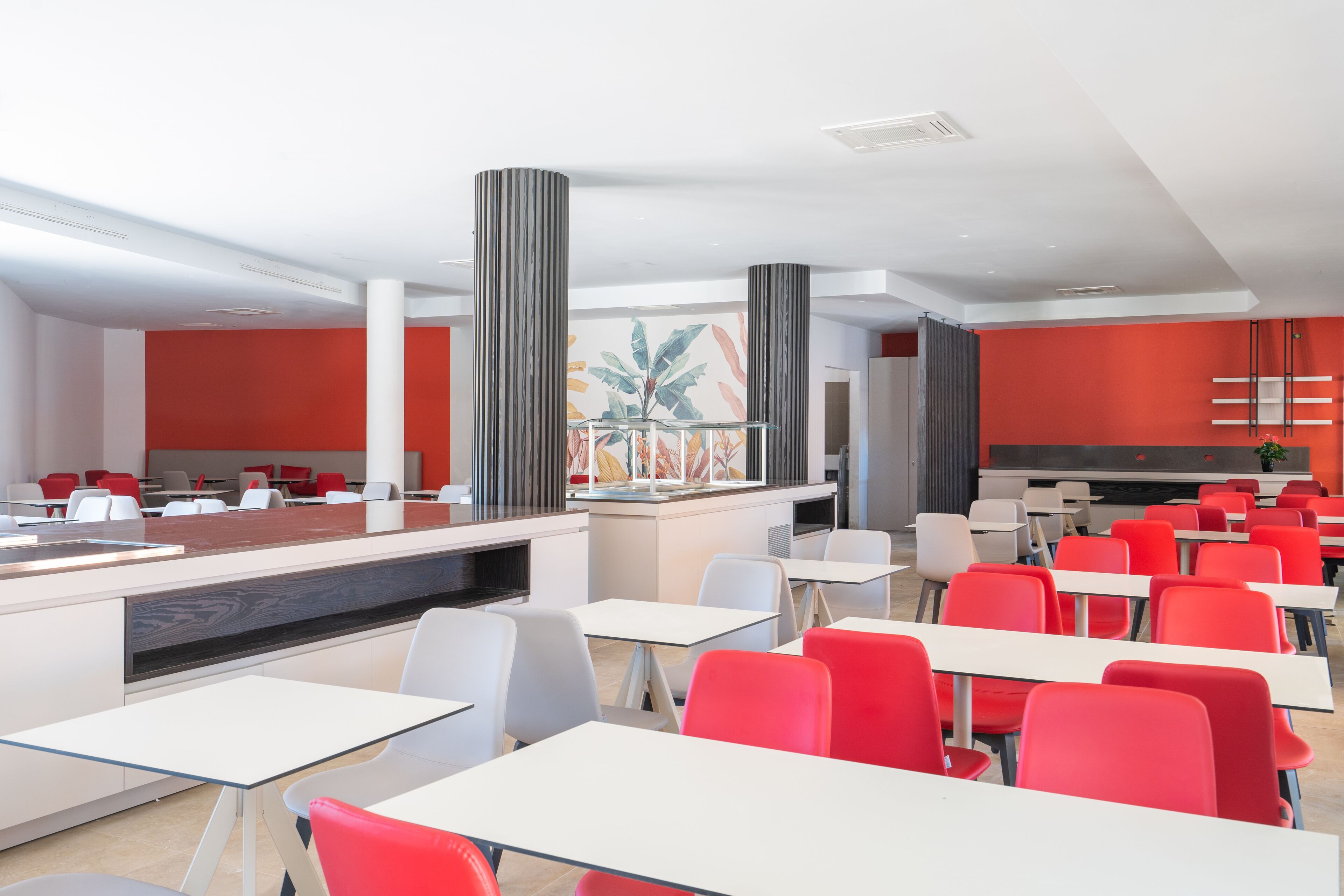 A modern dining area with white tables and red chairs, large pillars, and a bright red accent wall.