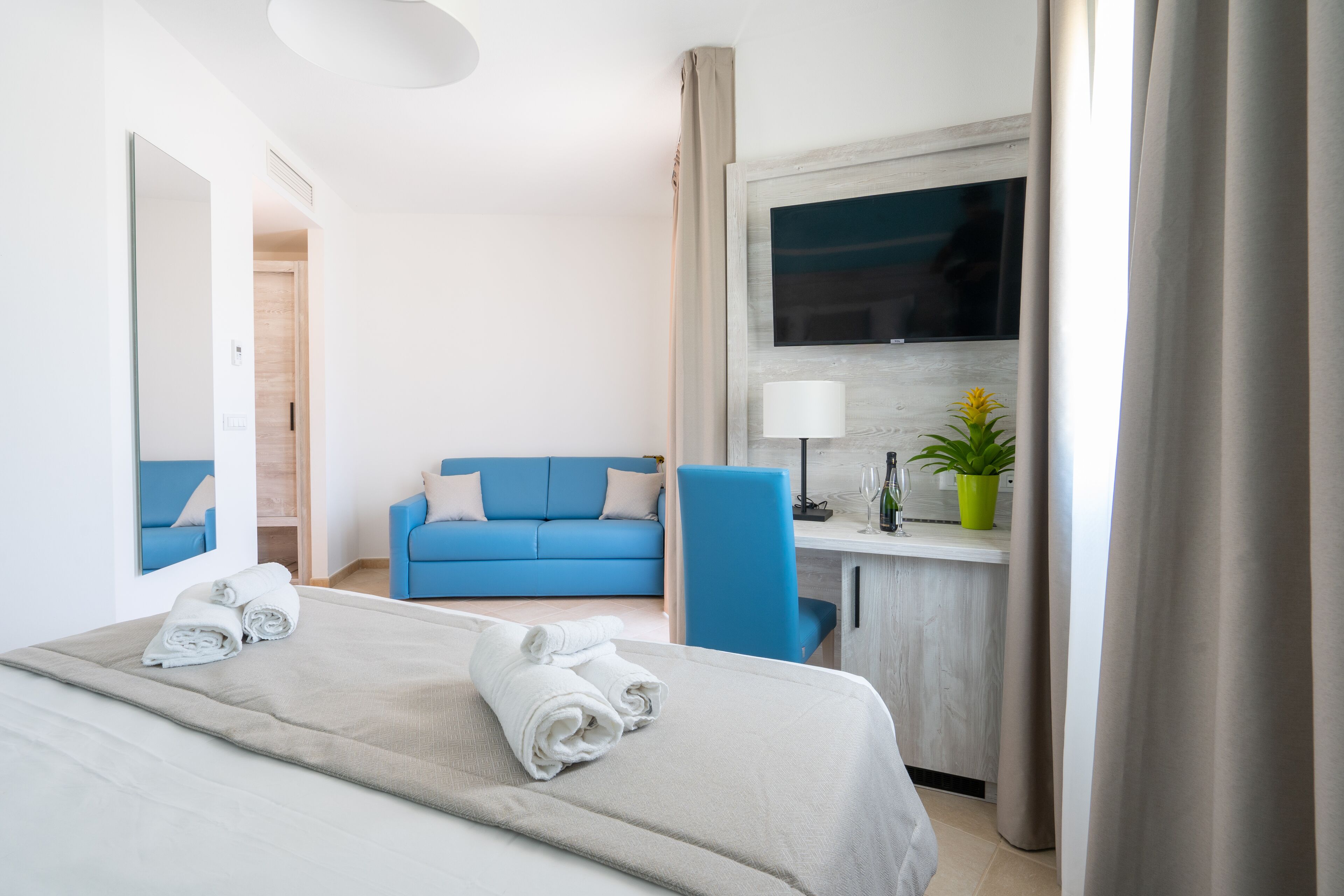 A hotel room with a bed, a blue sofa, a desk with a lamp, a TV, and a potted plant.