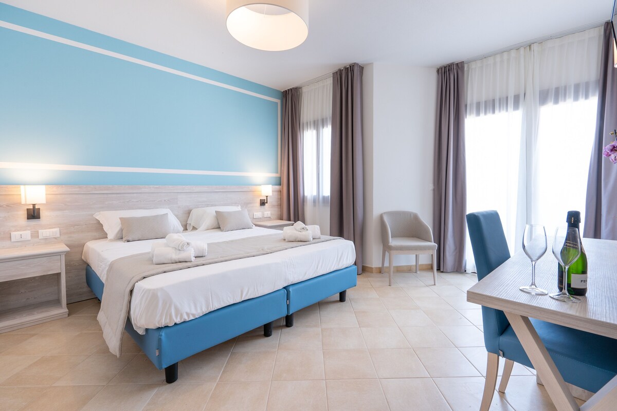 Une chambre d’hôtel avec un lit, des tables de chevet, une chaise, une table à manger avec des verres et une bouteille, et une grande fenêtre avec des rideaux.