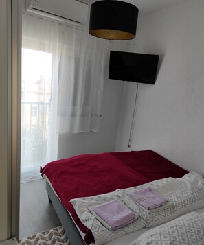 Une chambre à coucher avec un lit, une couverture rouge, un oreiller blanc, une serviette violette, un téléviseur noir fixé au mur et une fenêtre avec des rideaux légers.
