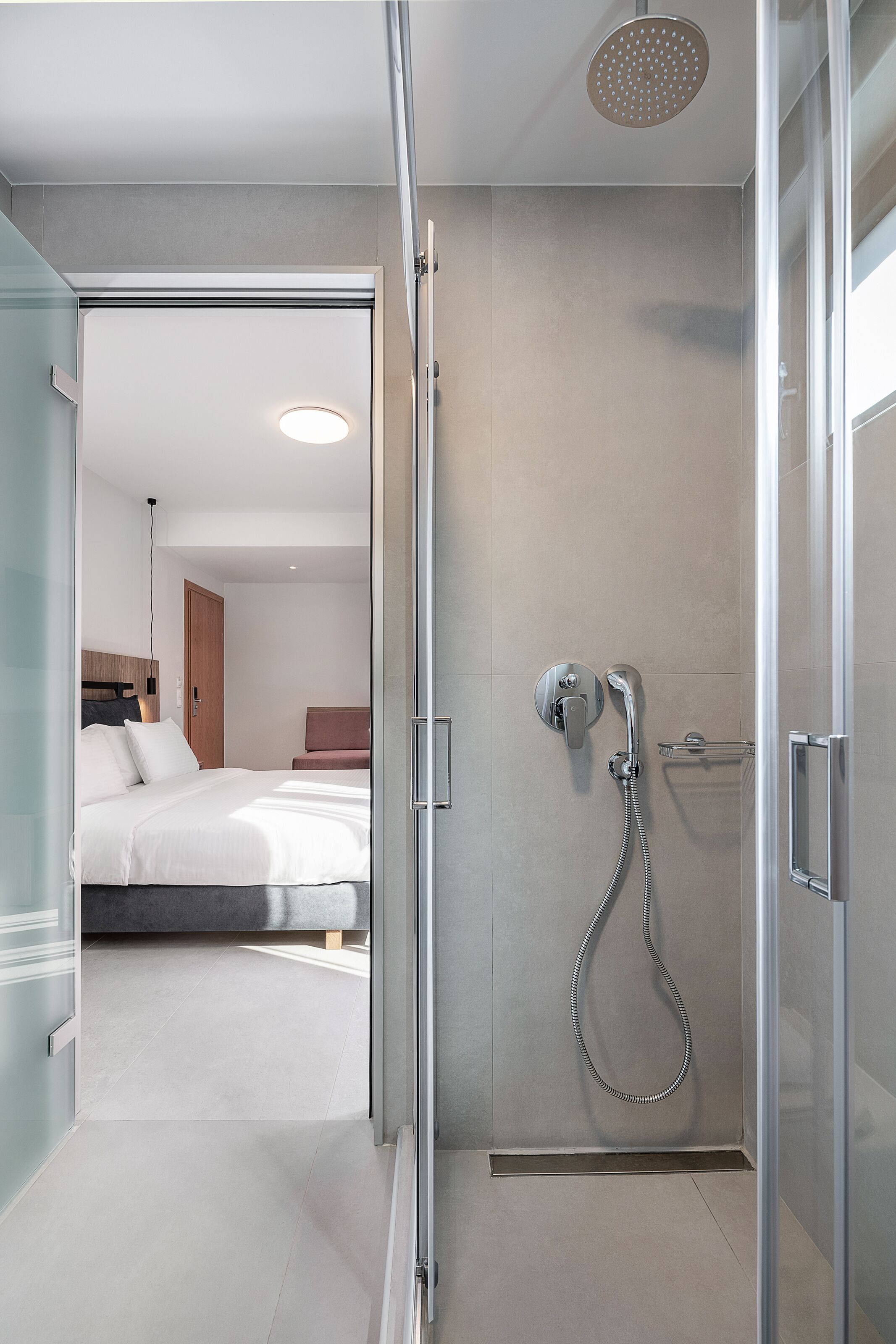 A modern hotel room with a glass shower enclosure, a bed with white linens, and a bedside table with a lamp.