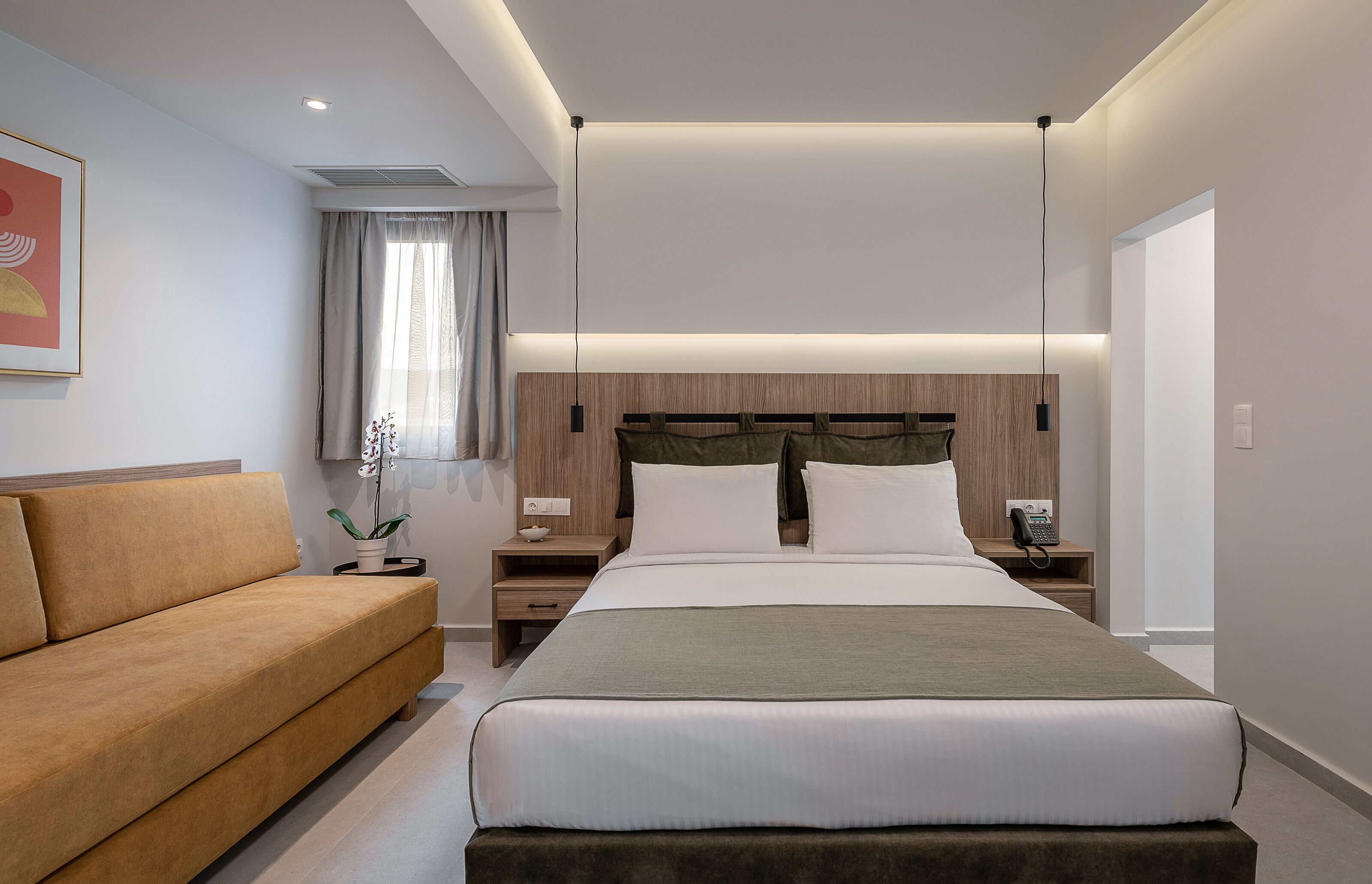 A modern hotel room with a large bed, a sofa, a bedside table with a phone, and a window with curtains.