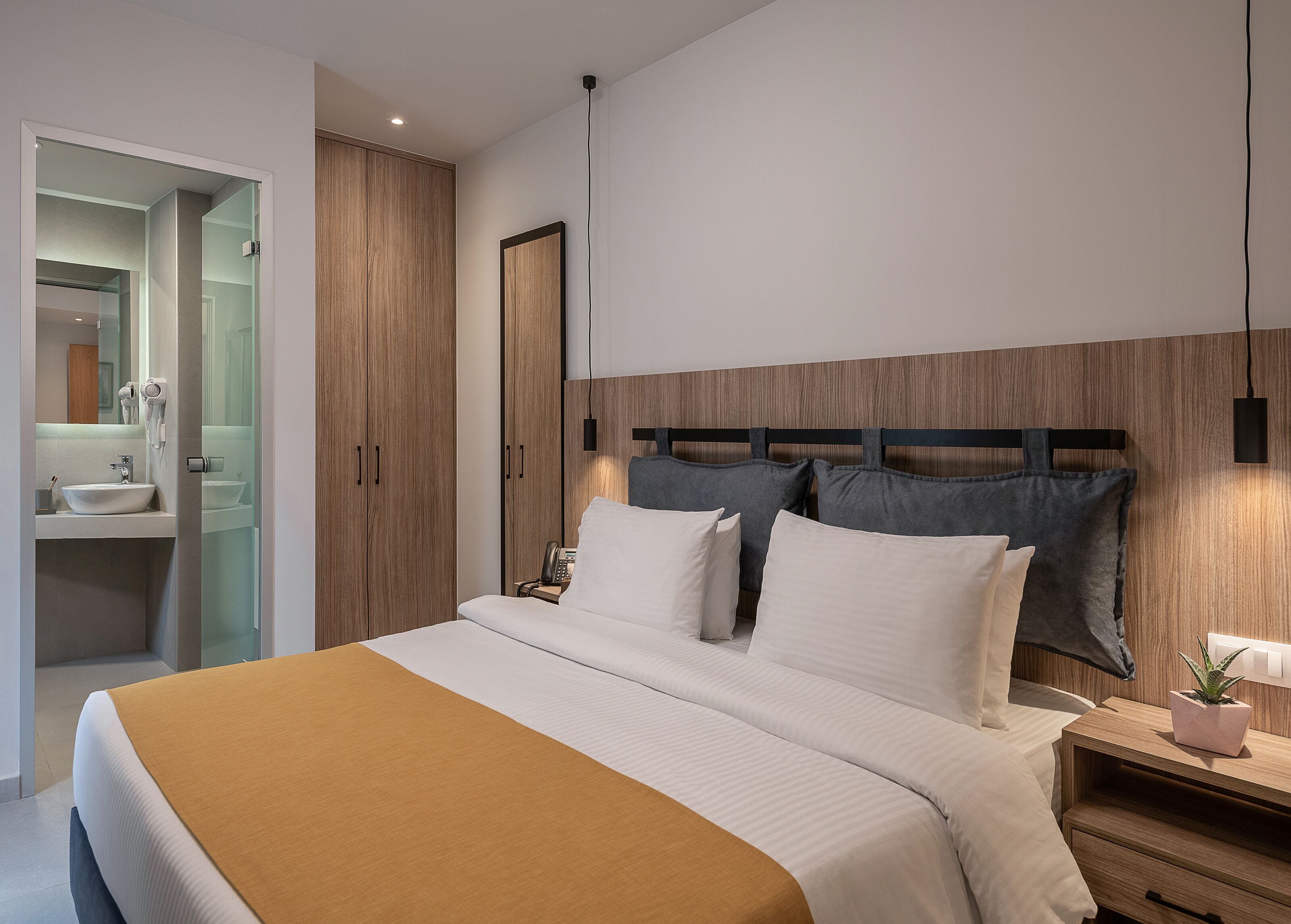 A modern hotel room with a large bed, a bedside table with a lamp, a small potted plant, and a wooden headboard.