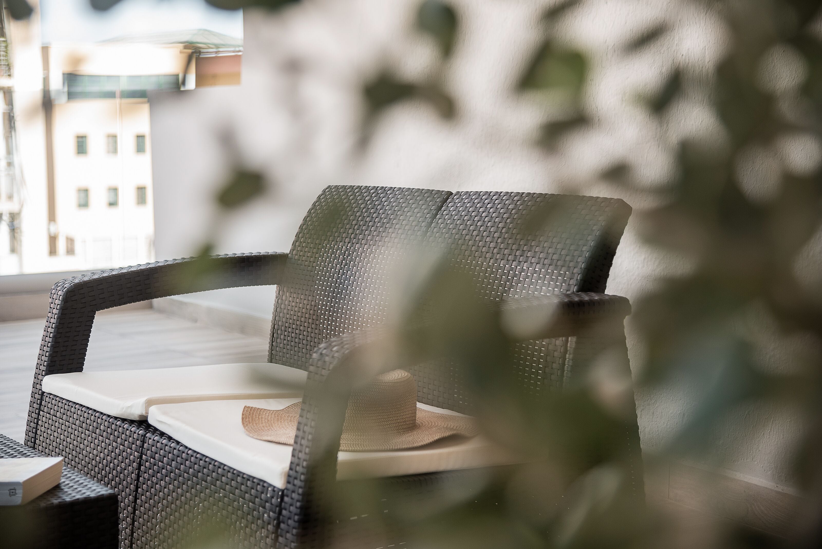 A wicker lounge chair with a white cushion and a woven hat placed on it, positioned near a window with a view of buildings.