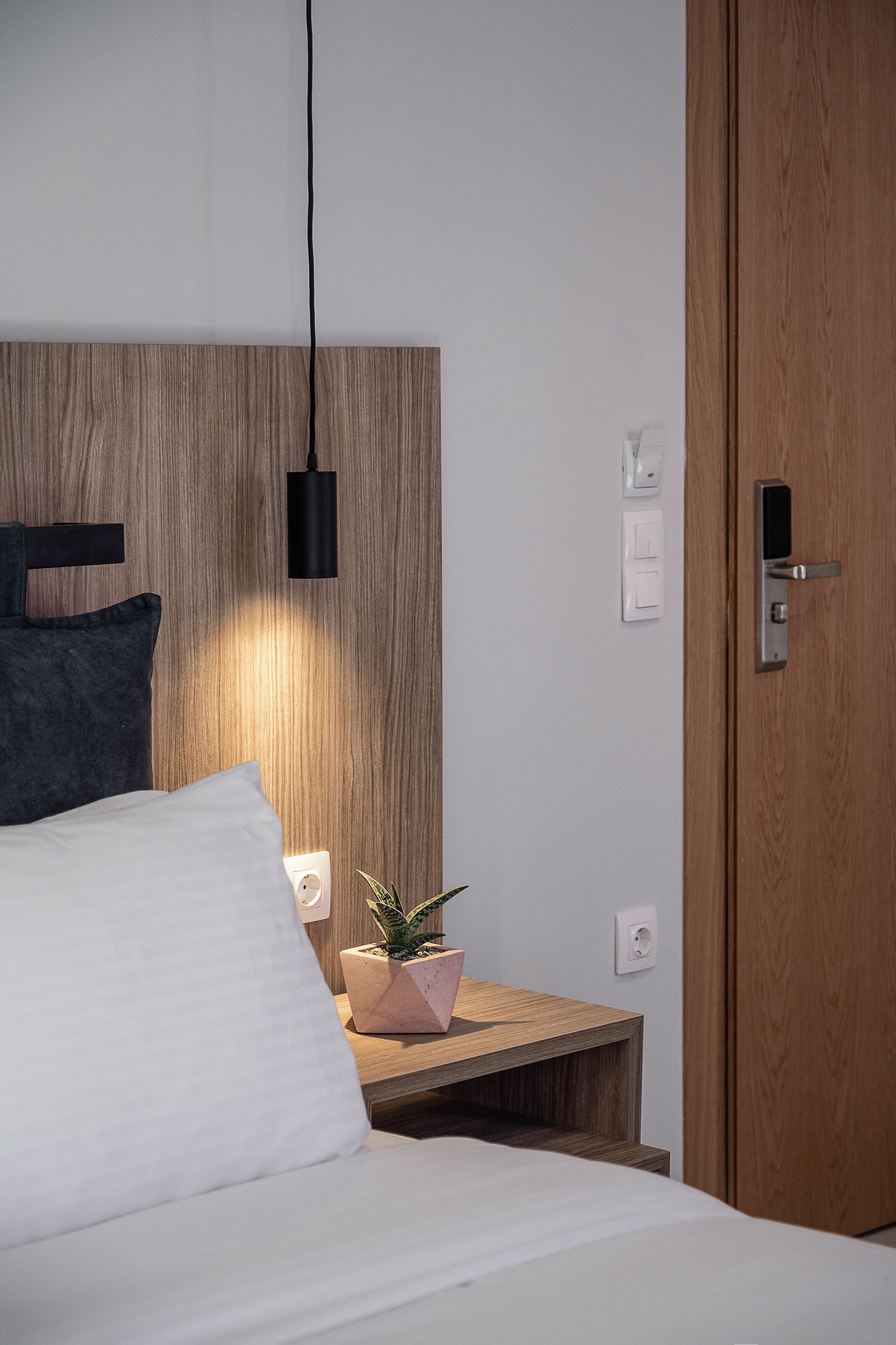 A neatly made bed with white linens, a wooden headboard, a bedside table with a pink plant, and a black pendant light.