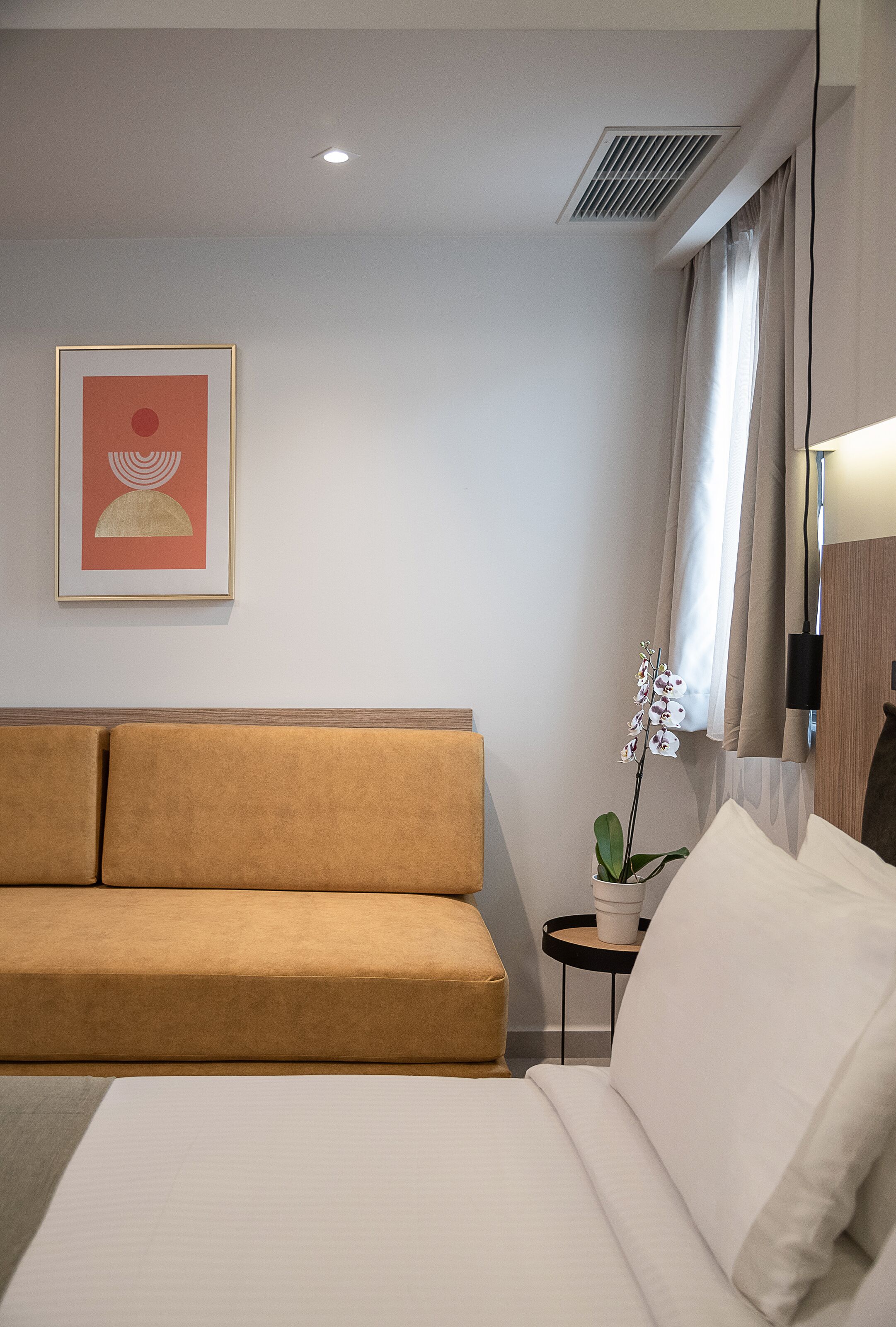 Una habitación de hotel moderna con un sofá color beige, una cama blanca, una obra de arte enmarcada en la pared, una orquídea en maceta y una mesita auxiliar pequeña.