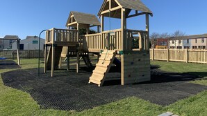 Un parque infantil de madera con estructuras para escalar y un tobogán, rodeado por una cerca y un campo cubierto de césped.