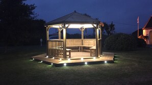Un quiosco de madera con un banco y una mesa, rodeado por una cerca baja e iluminado por luces en el suelo.