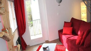 A living room with a red sofa, a white coffee table, and a red rug. There is a window with red curtains, a wall-mounted light fixture, and a wooden shelf with books.