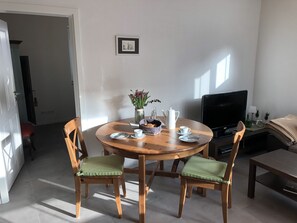 A dining area with a round wooden table, two chairs with green cushions, a vase with flowers, a coffee pot, and a television on a stand.