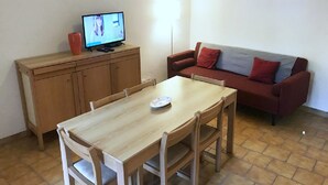 Ett litet, möblerat rum med ett träbord och stolar, en röd soffa, en tv på ett träskåp och en lampa.
