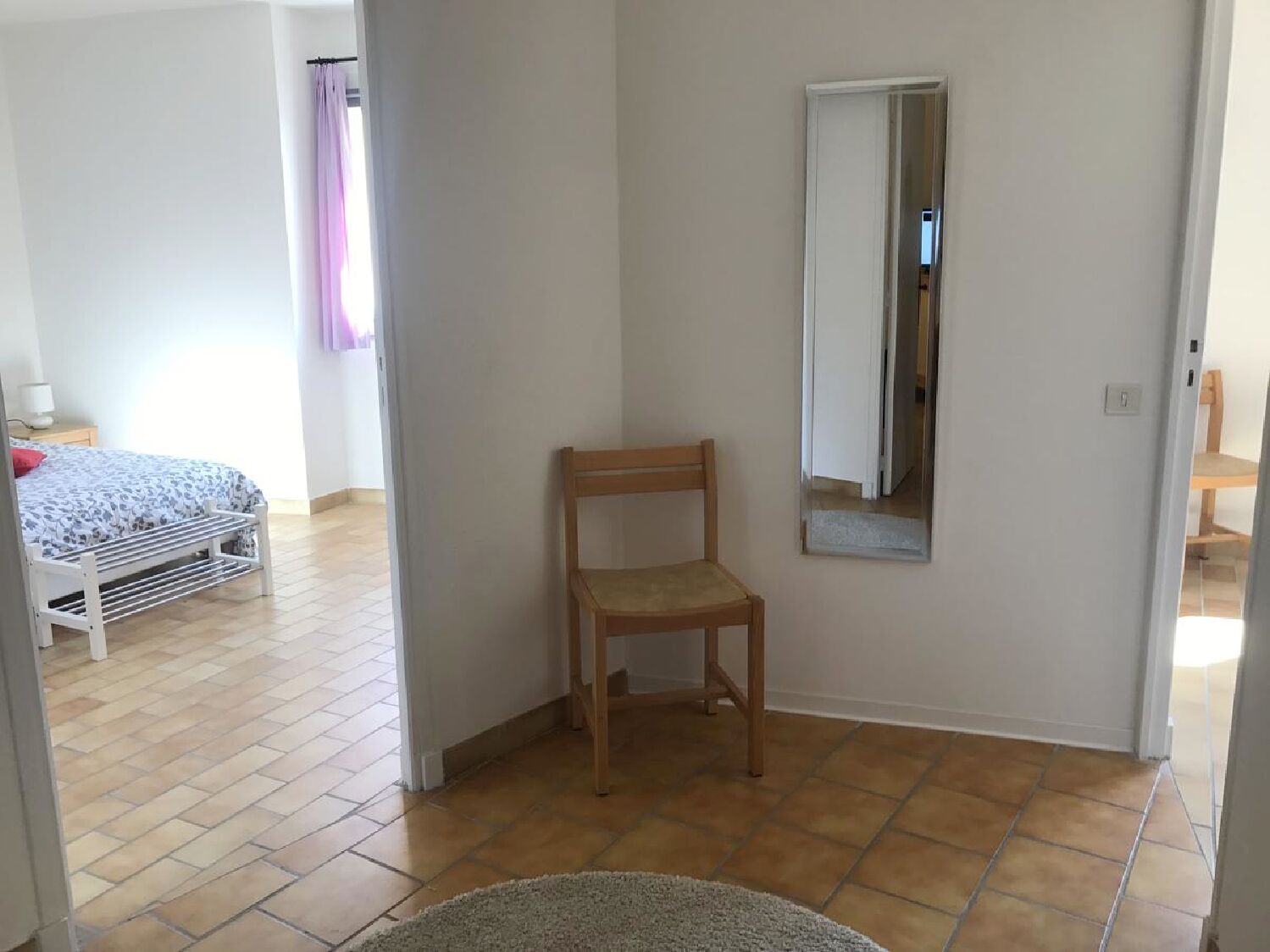 A room with a bed, a chair, a mirror, and a tiled floor.