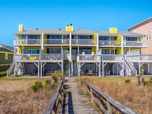 A multi-story building with balconies, a wooden boardwalk leading to the entrance, and a clear blue sky.