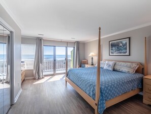 A bedroom with a four-poster bed, a balcony with a view of the beach, and a painting on the wall.
